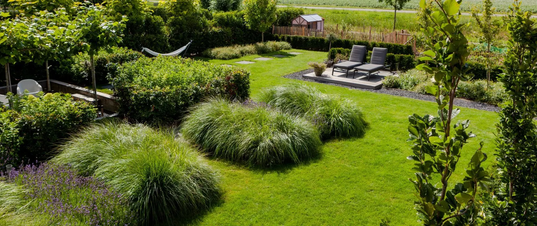 Verzorgde tuin met gazon, siergrasplanten, lavendel, ligbedden op terras, hangmat en tuinhuisje op de achtergrond