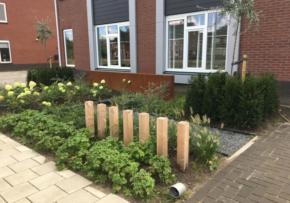 Voortuin met beplanting, houten palen, siergrassen en een cortenstalen scherm voor een bakstenen woning
