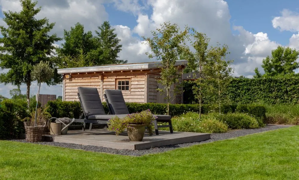Twee ligstoelen op een stenen terras voor een houten tuinhuis, omgeven door groen gazon en beplanting