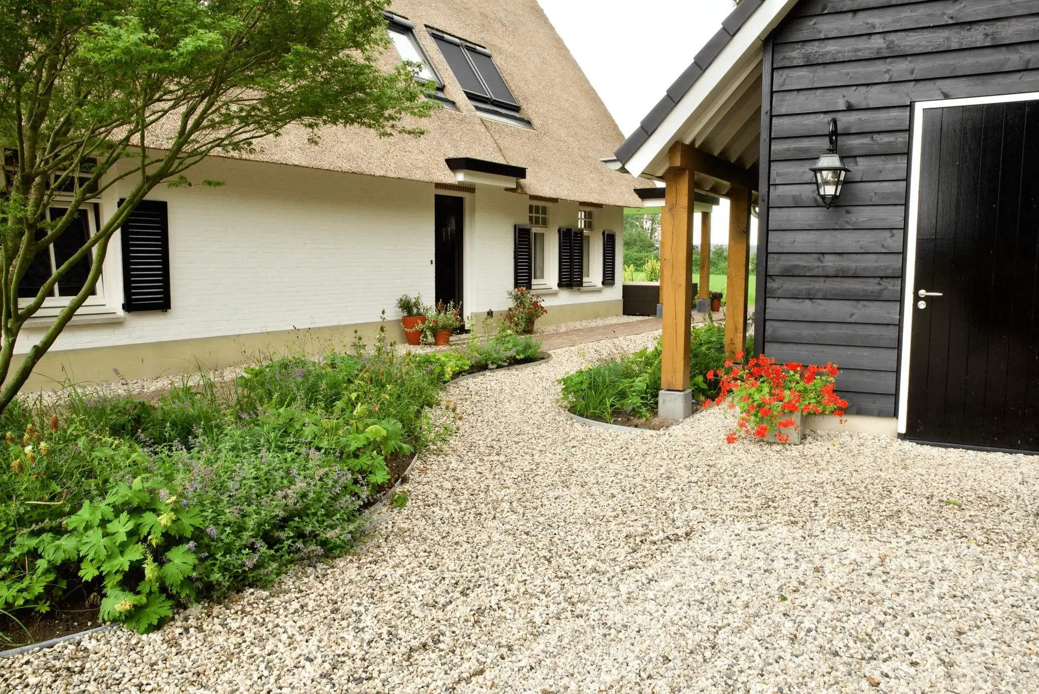 Voortuin van een rietgedekte woning met grindpad, beplantingsborders en een zwarte houten bijgebouw met overkapping