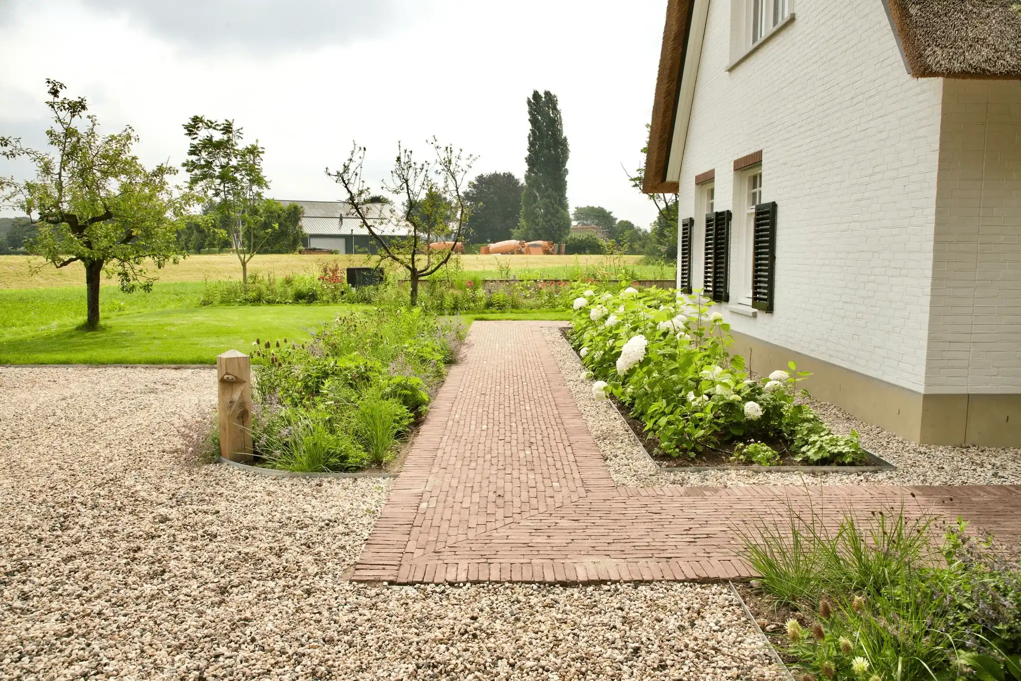 Bakstenen pad langs een rietgedekte boerderij met borders van hortensia's, grind en fruitbomen in een landelijke tuin