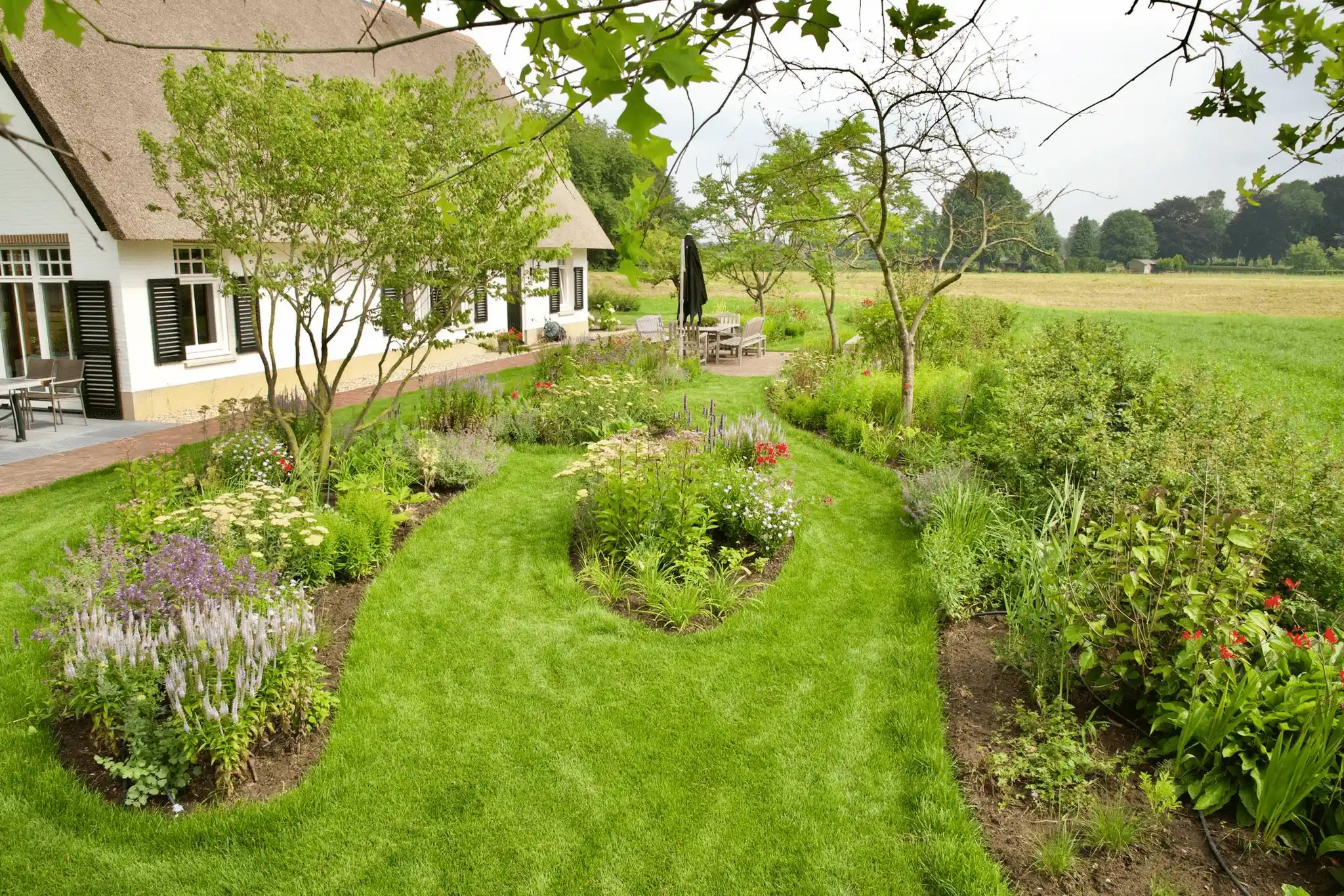 Landelijke tuin met golvende grasvelden, kleurrijke borders en fruitbomen naast een wit rieten huis met uitzicht op weilanden