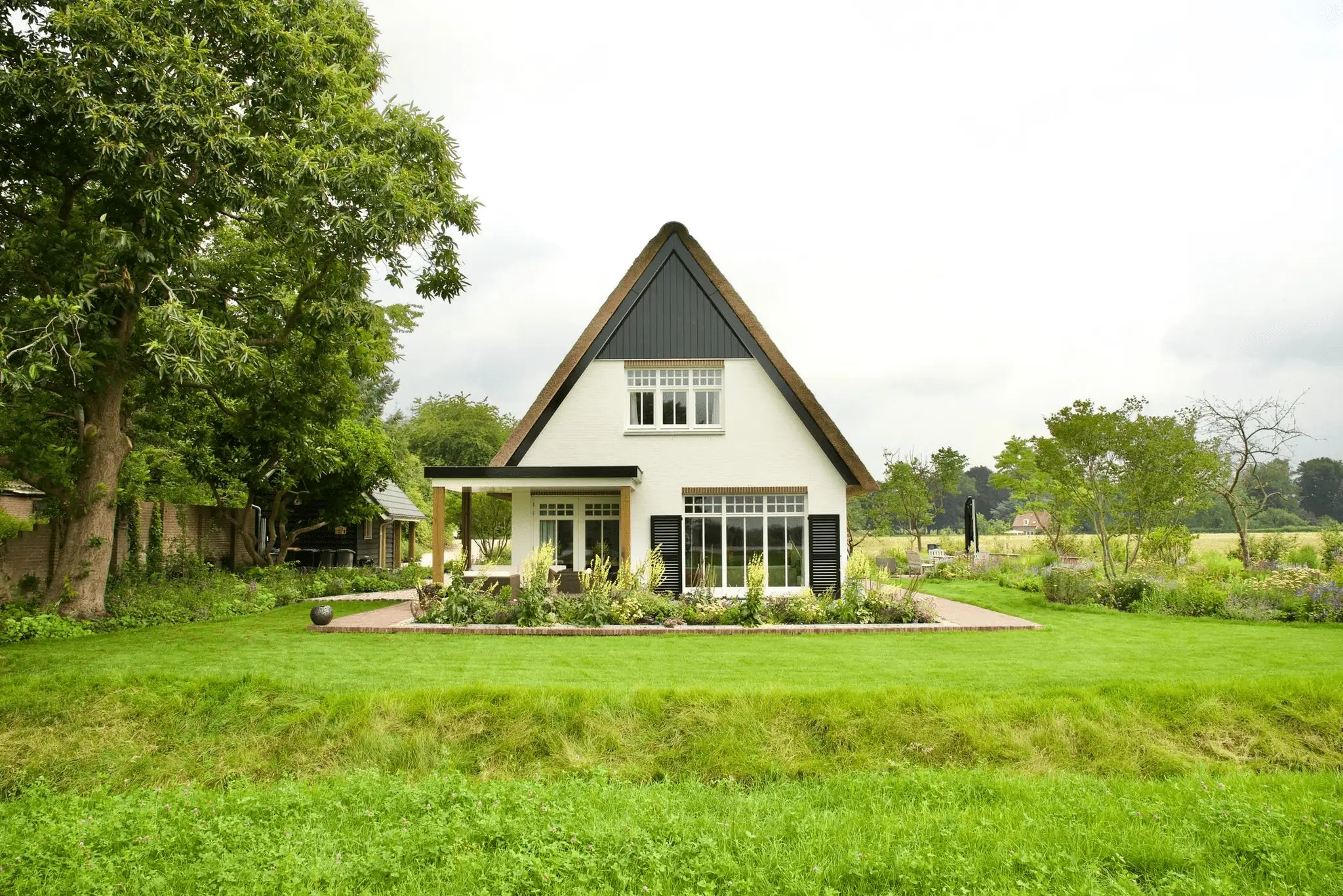 Wit rieten huis met puntig dak, omgeven door een weelderige tuin en open landschap op de achtergrond