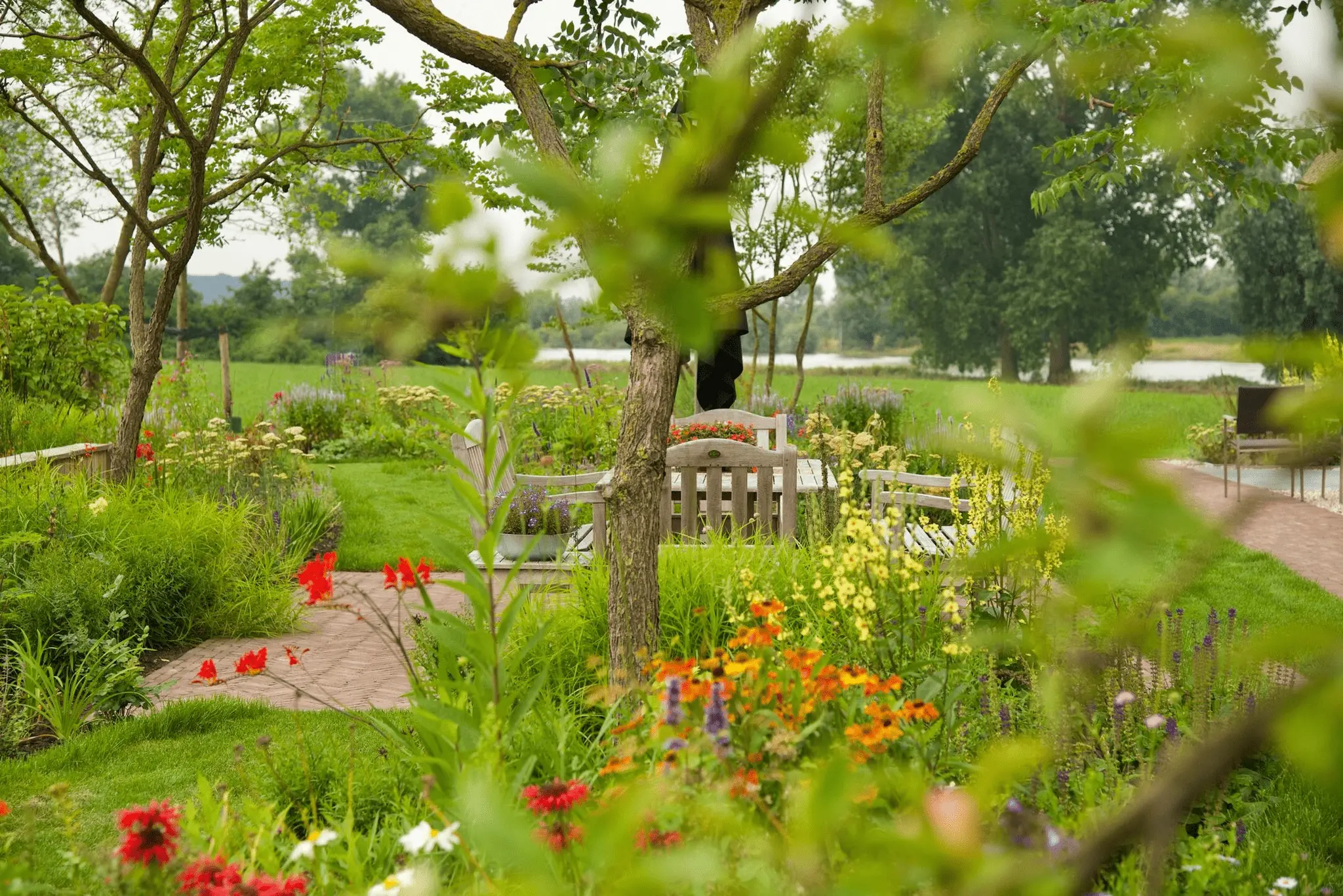 Weelderige tuin met kleurrijke bloemen, bomen en houten tuinstoelen, met een waterpartij op de achtergrond