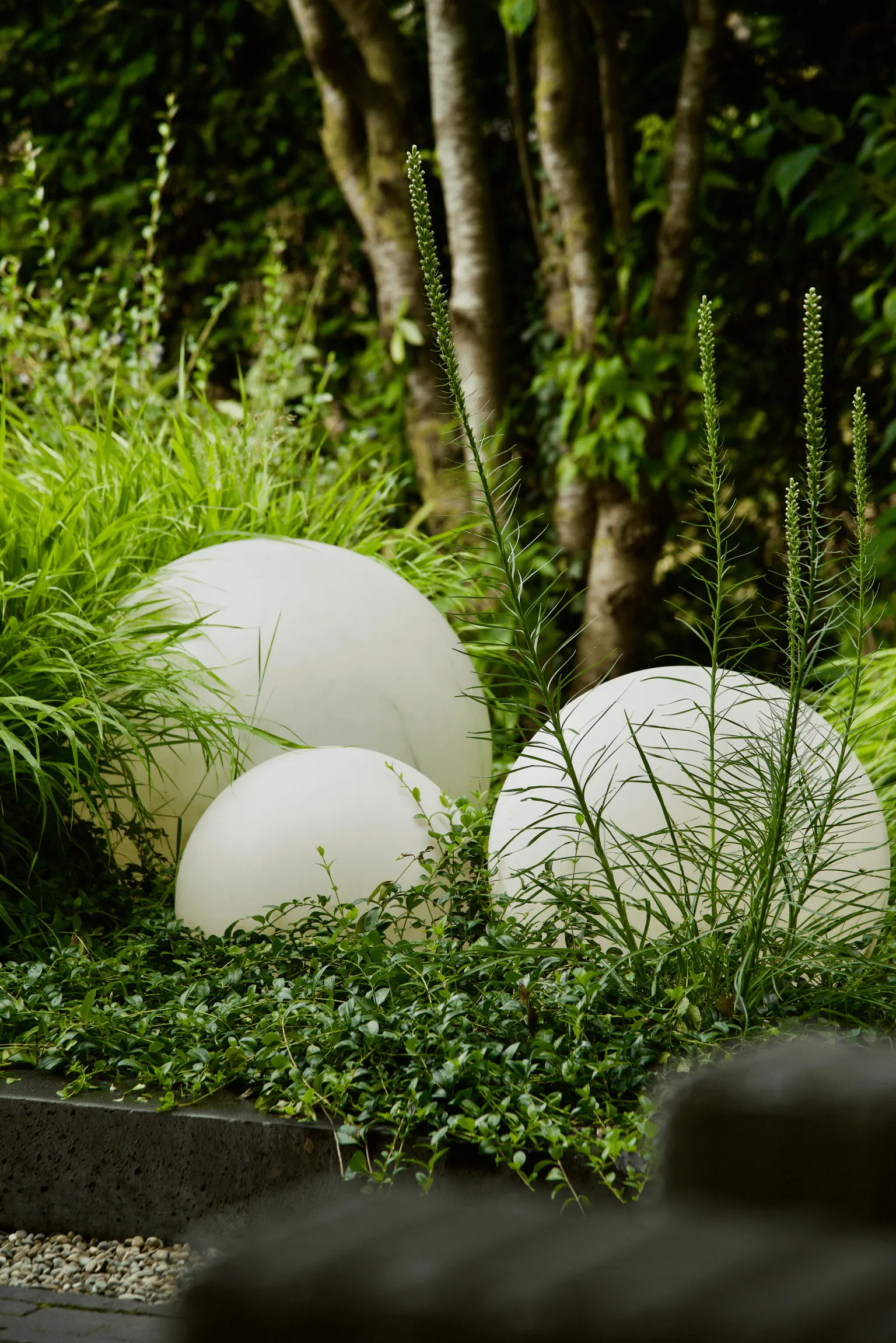 Drie witte ronde buitenlampen in verschillende groottes tussen groen tuinbeplanting en siergras