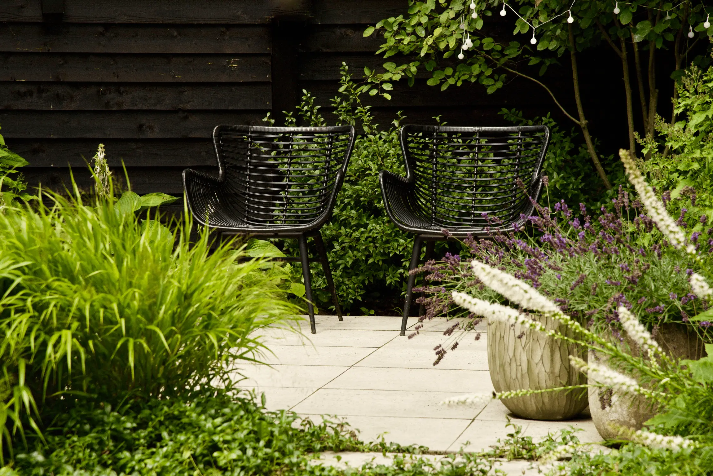 Twee zwarte rotan tuinstoelen op een licht terras, omgeven door weelderig groen, lavendel en witte bloemen in stenen potten