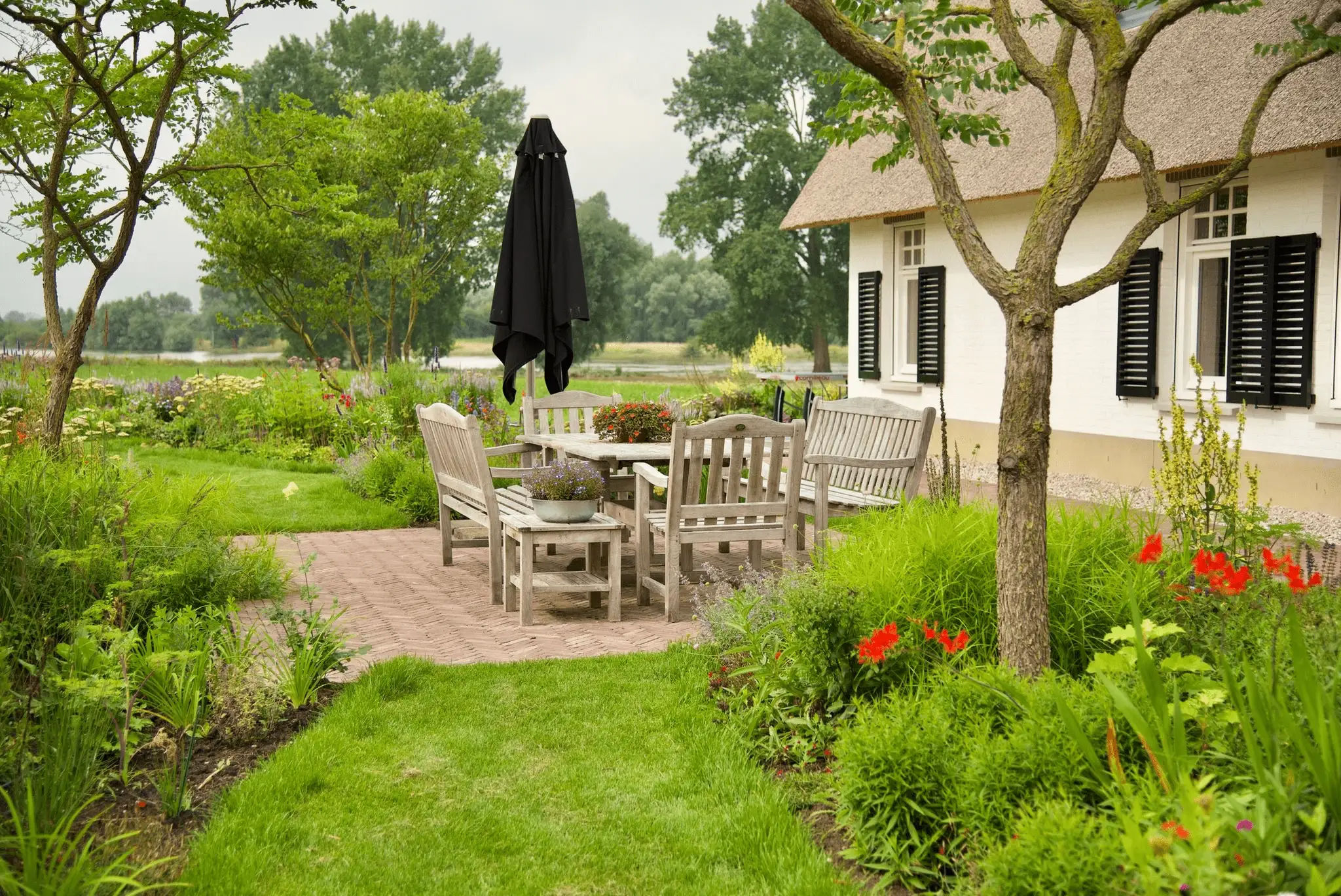 Landelijke tuin met houten tuinmeubelen op een terras naast een rieten woning, omgeven door bloeiende borders en bomen
