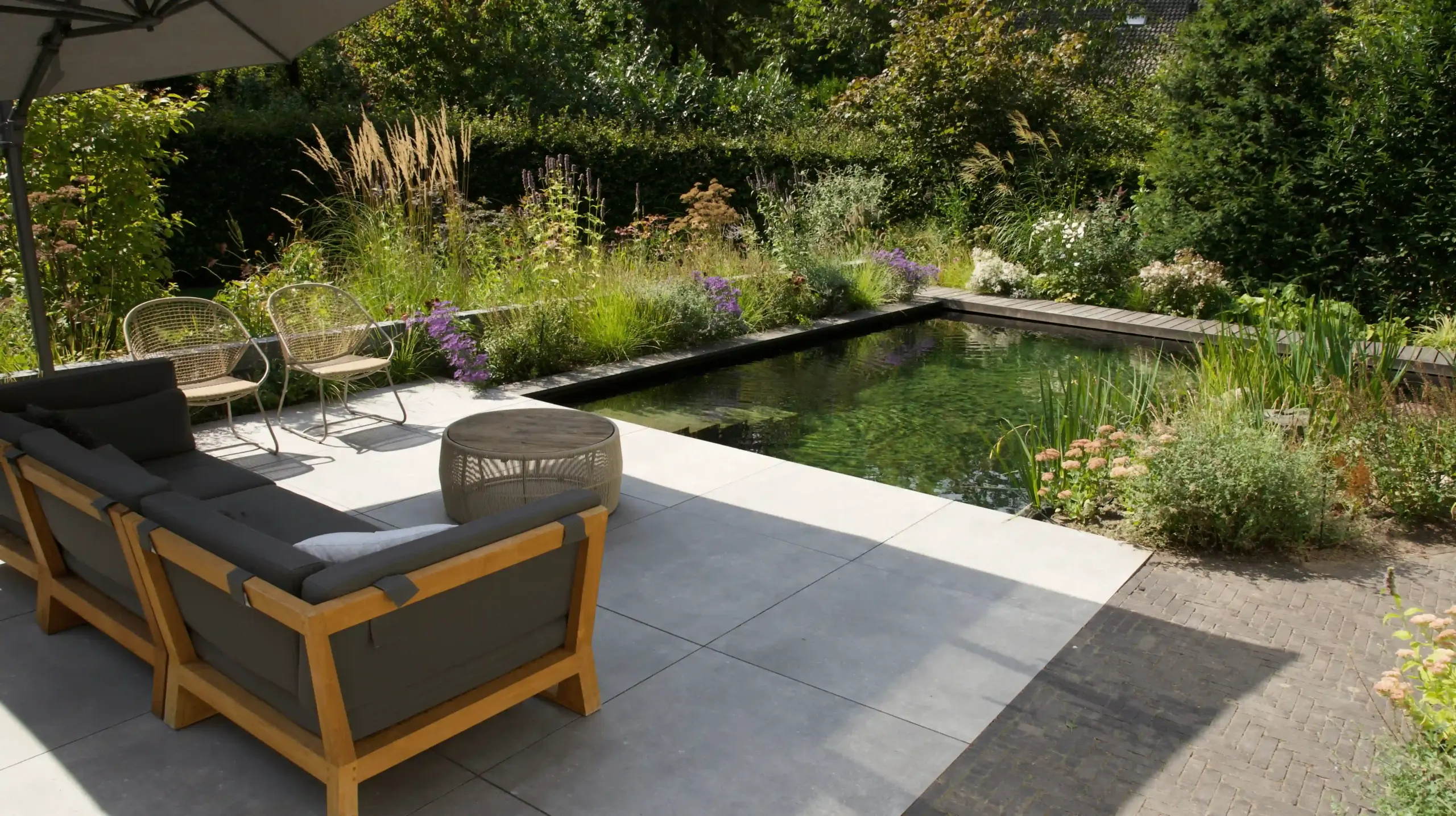 Moderne tuin met een natuurlijk zwembad, omgeven door weelderige beplanting en een terras met houten tuinmeubelen en parasol