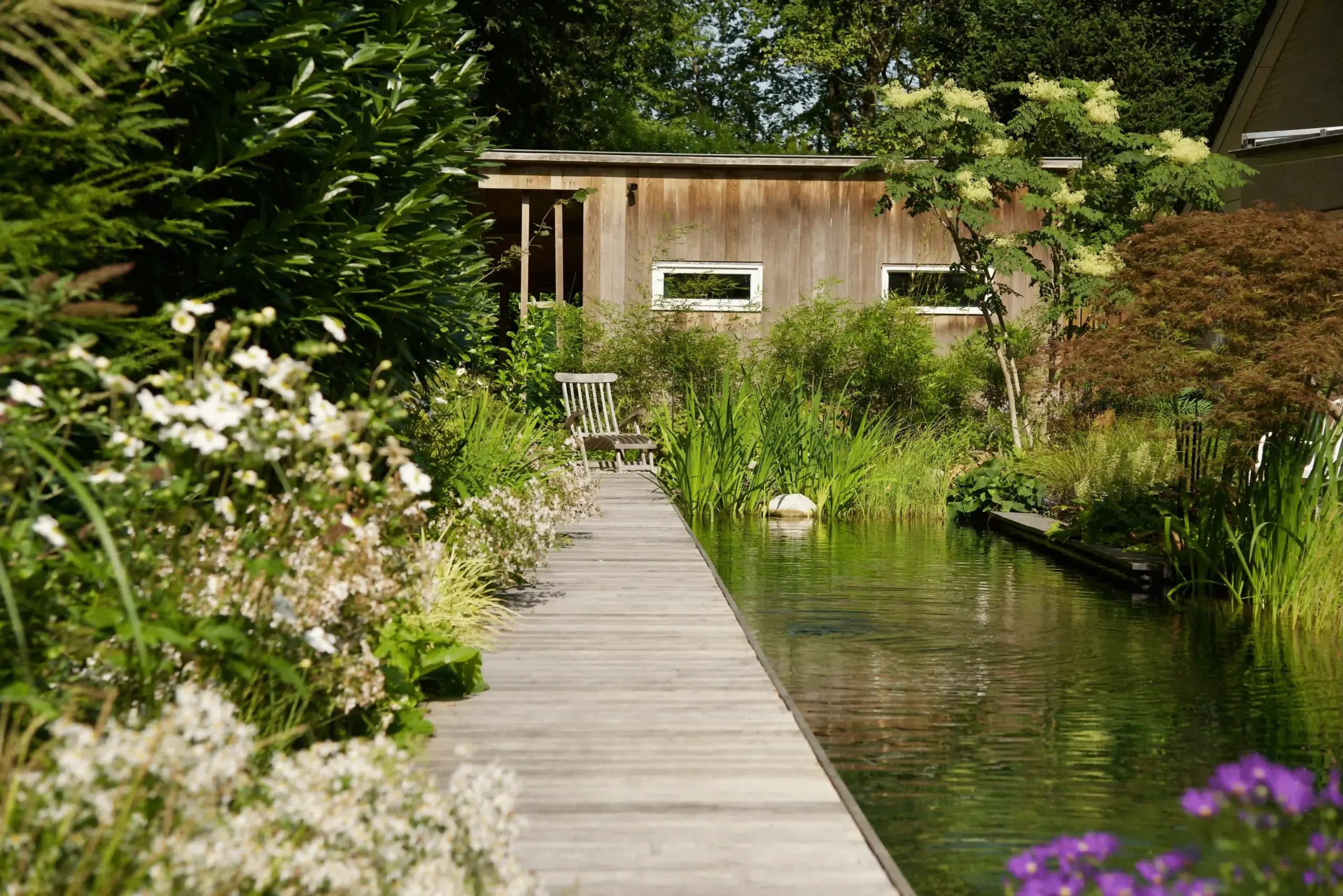 Houten vlonder langs een natuurlijke vijver met weelderige beplanting en een tuinstoel, met op de achtergrond een houten tuin
