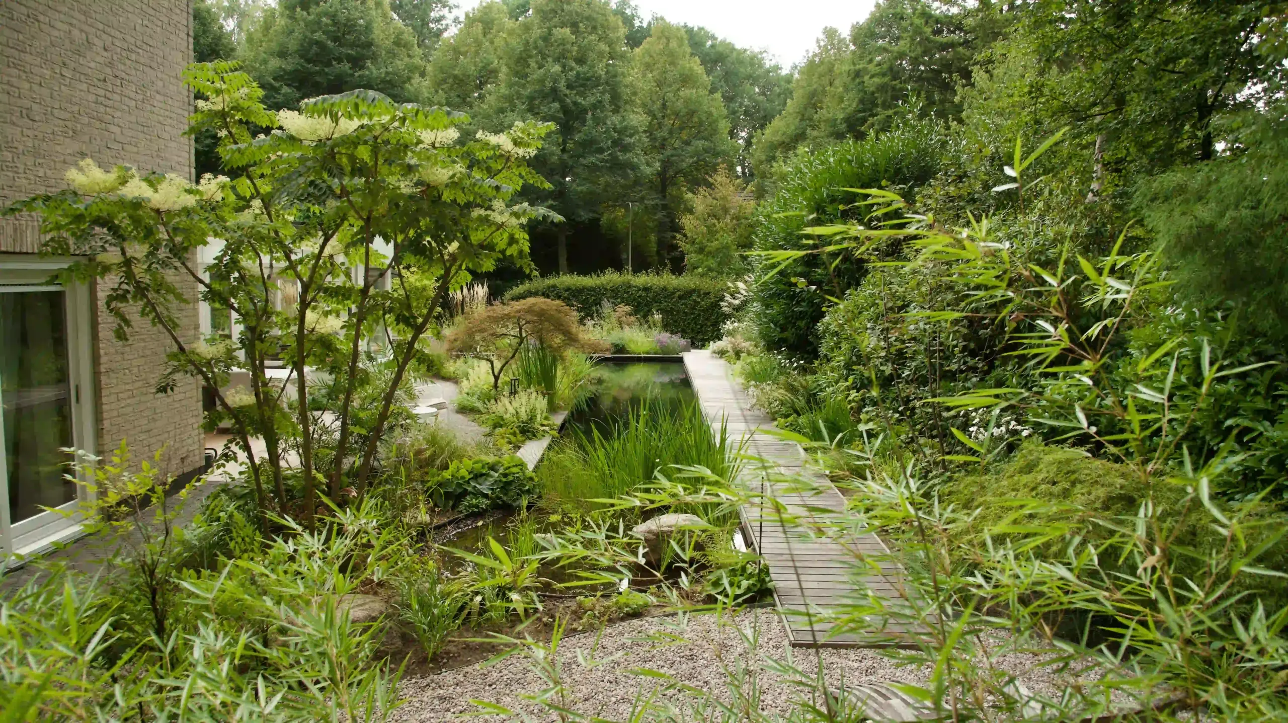 Weelderige tuin met vijver, houten vlonder, grindpad en gevarieerde beplanting naast een woning