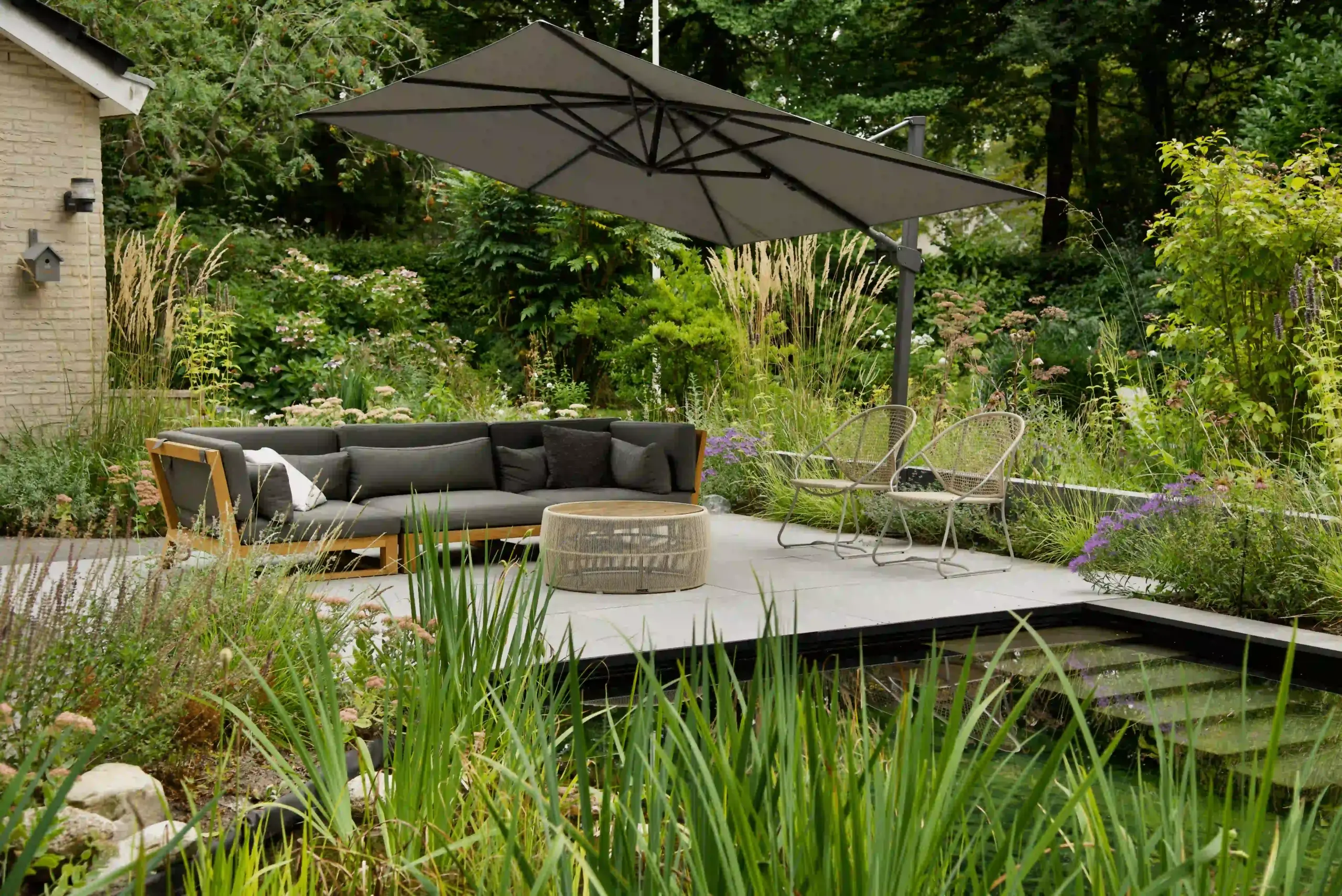 Moderne tuinterras met hoekbank, loungestoelen en een grote zweefparasol, omgeven door weelderige beplanting en een vijver