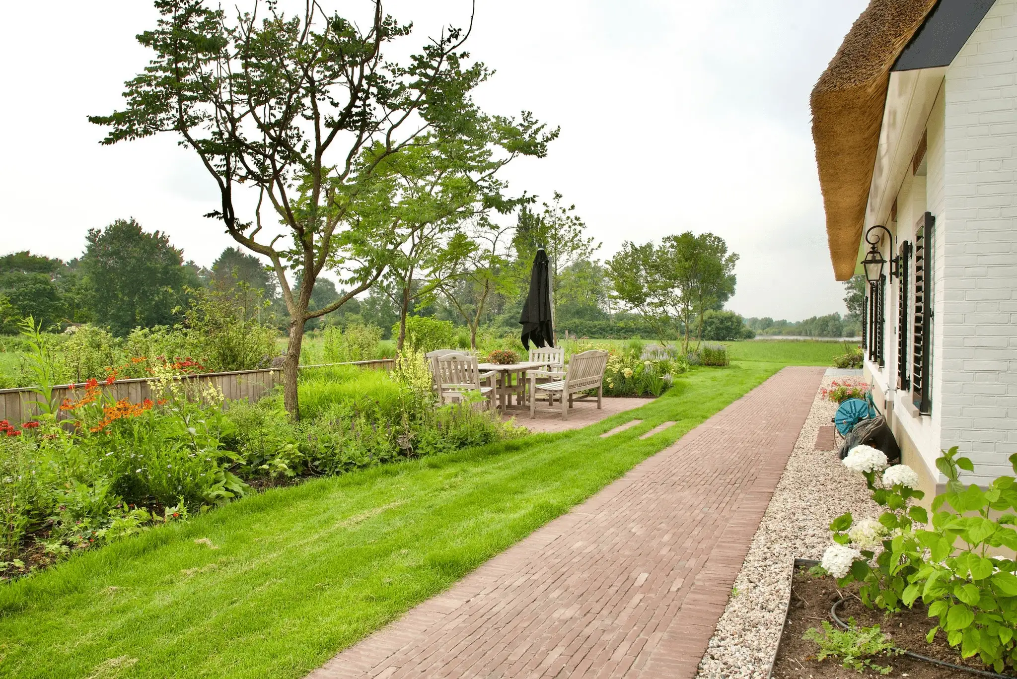 Tuin van een wit rieten huis met bakstenen pad, houten tuinset, bloemborders en uitzicht op het open landschap
