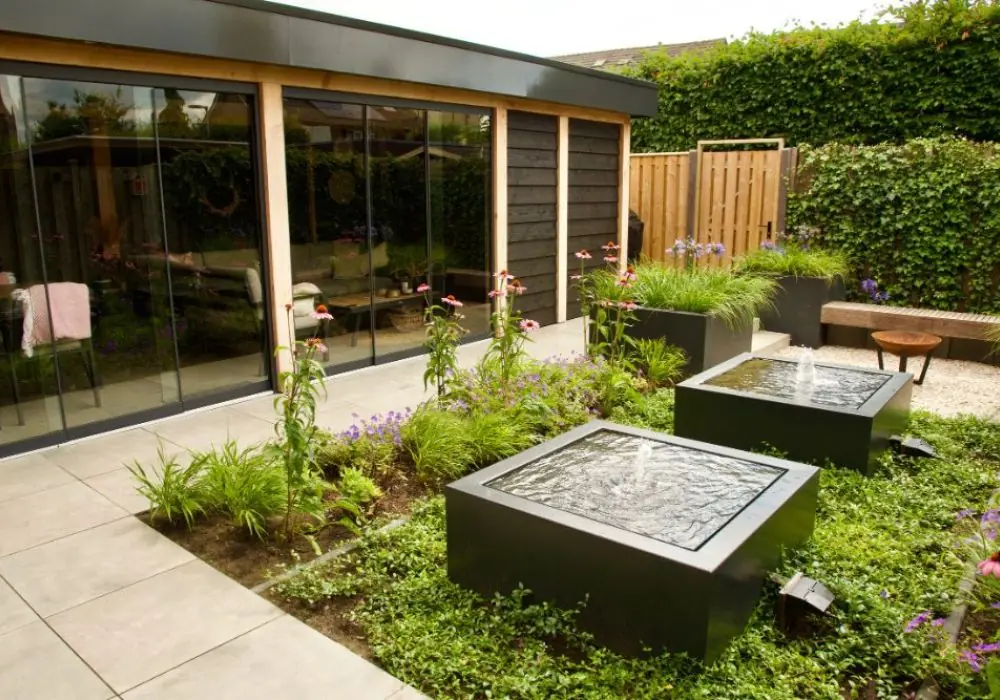 Moderne tuin met twee vierkante waterornamentenbakken, kleurrijke beplanting en een stijlvol tuinhuis met glazen schuifpuien