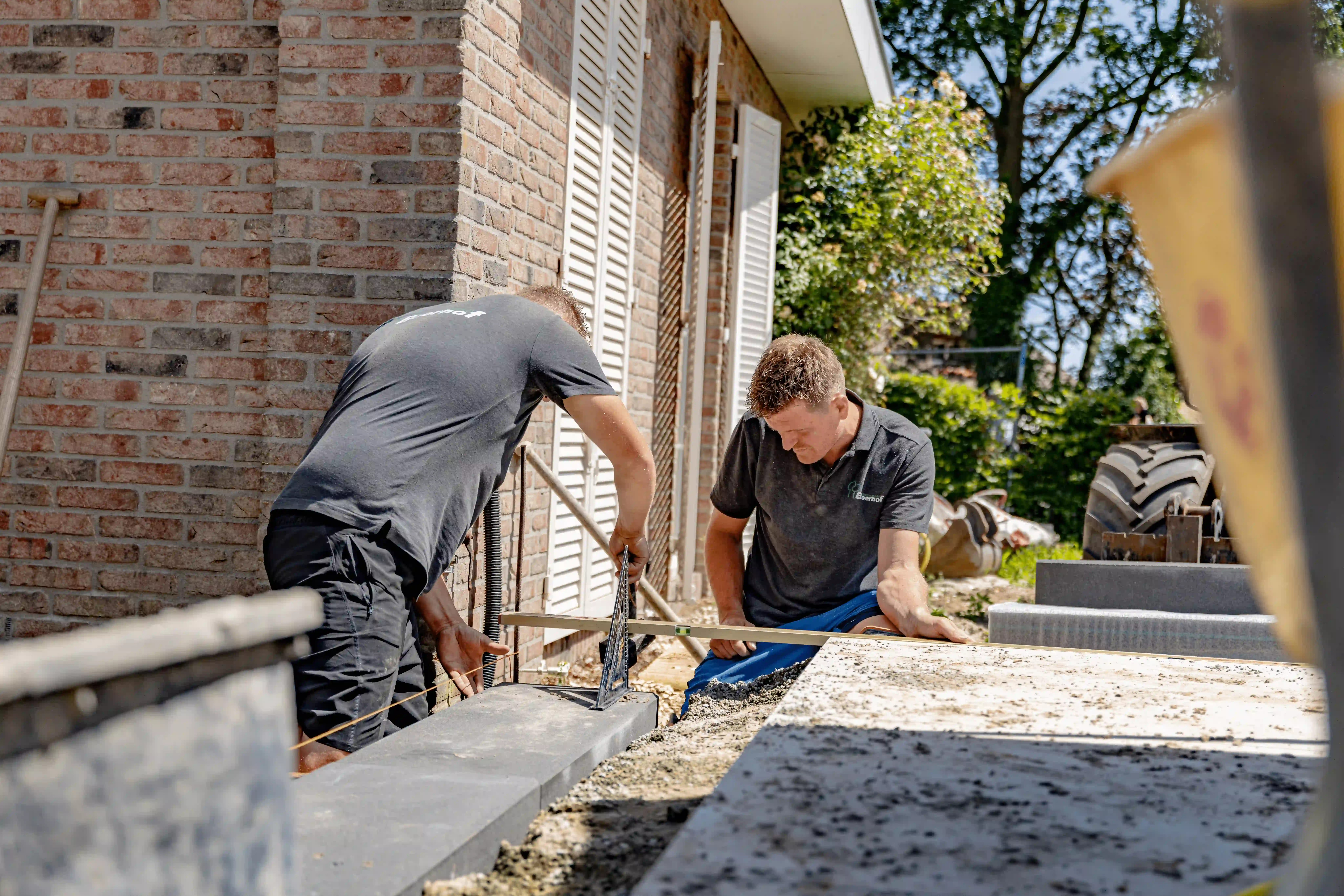 Twee werklieden plaatsen betonnen terrastegels met waterpas naast een bakstenen woning