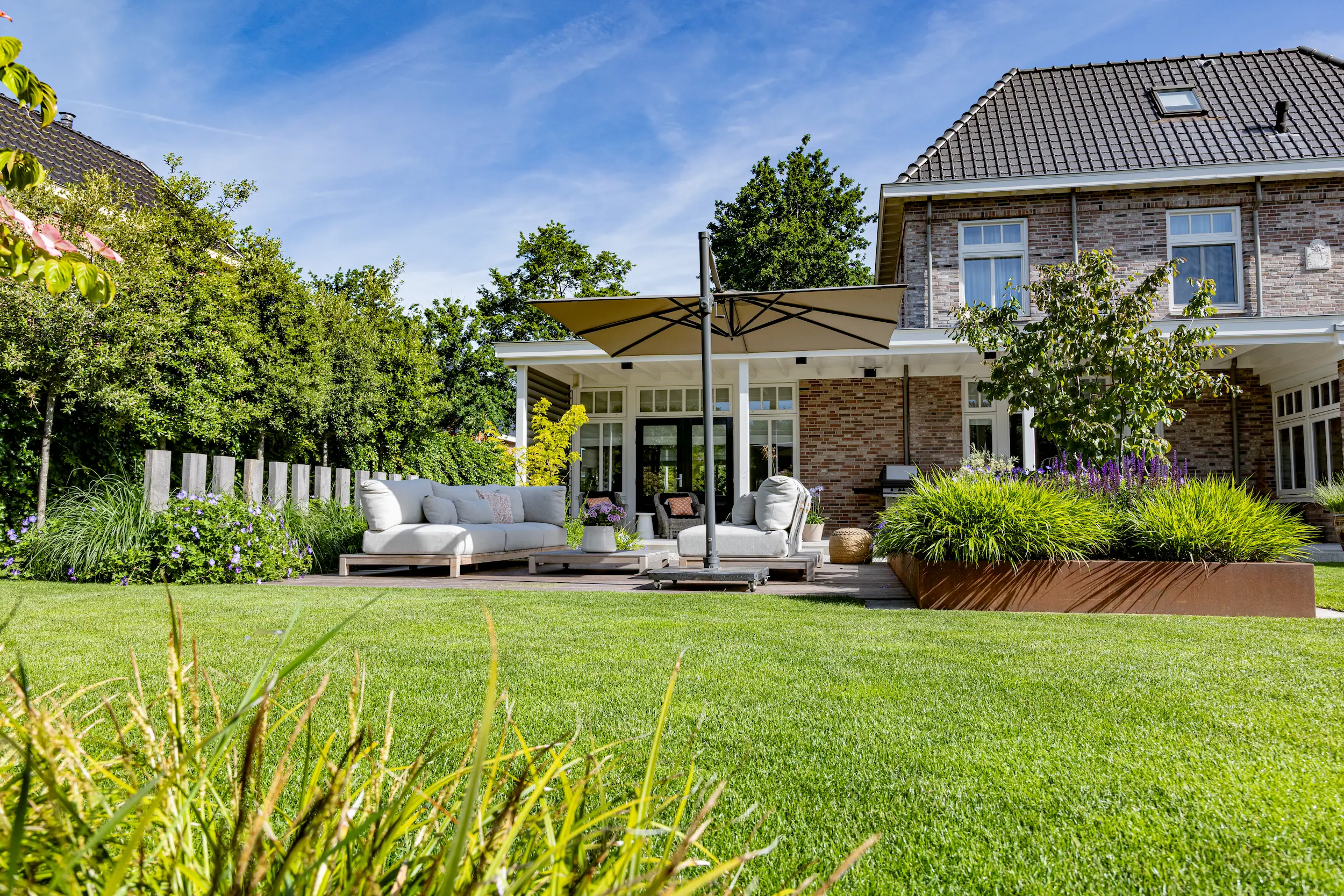 Strak gazon voor een houten terras met grijze loungebanken en zweefparasol bij een bakstenen villa met cortenstalen borders.