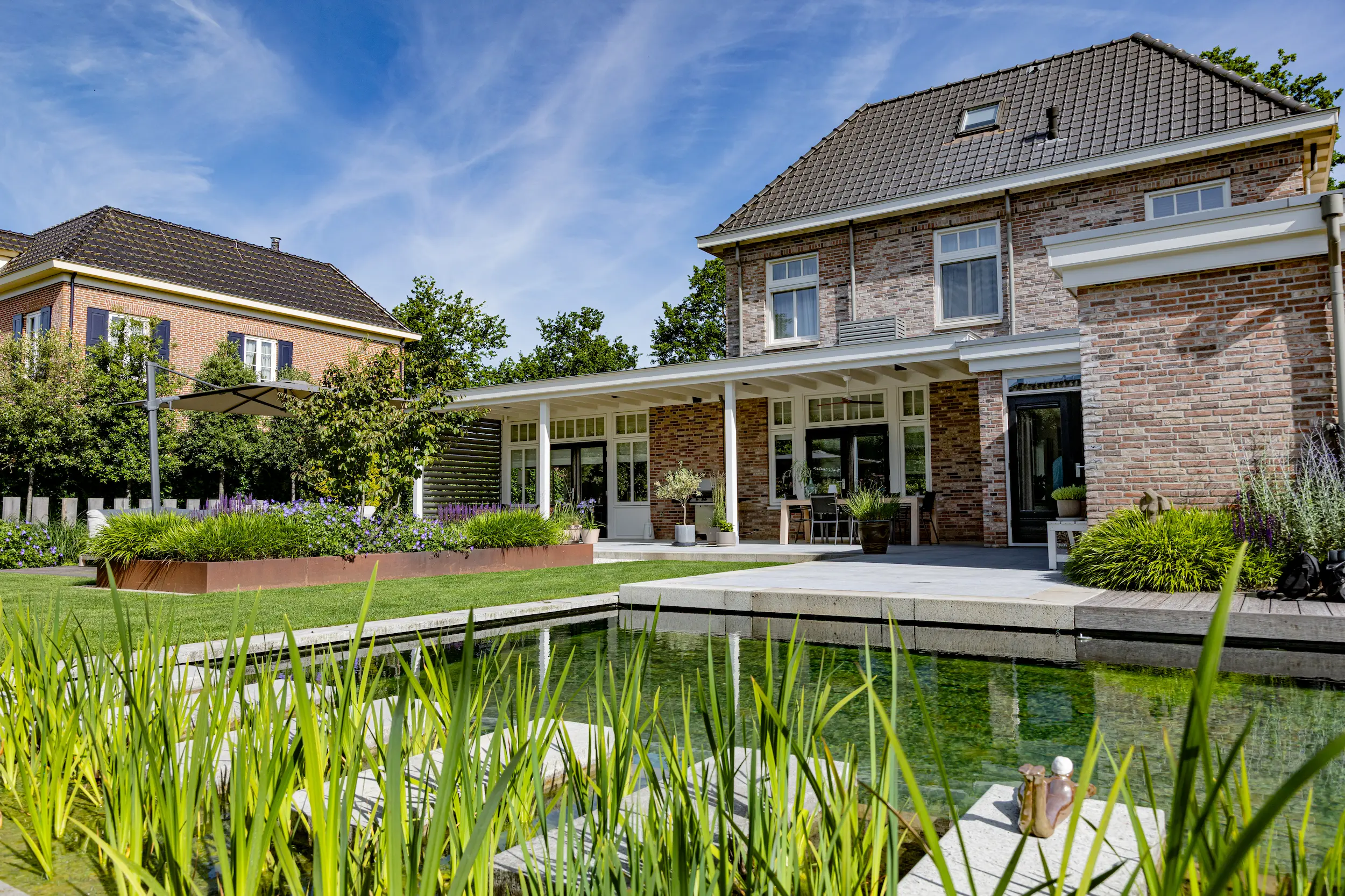Bakstenen woning met witte veranda, terras en natuurlijke zwemvijver omringd door grassen en cortenstalen plantenbakken.