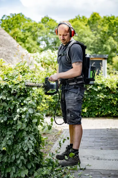 Man met gehoorbescherming en rugzakaccu snoeit een beukenhaag met een elektrische heggenschaar op een tuinpad.