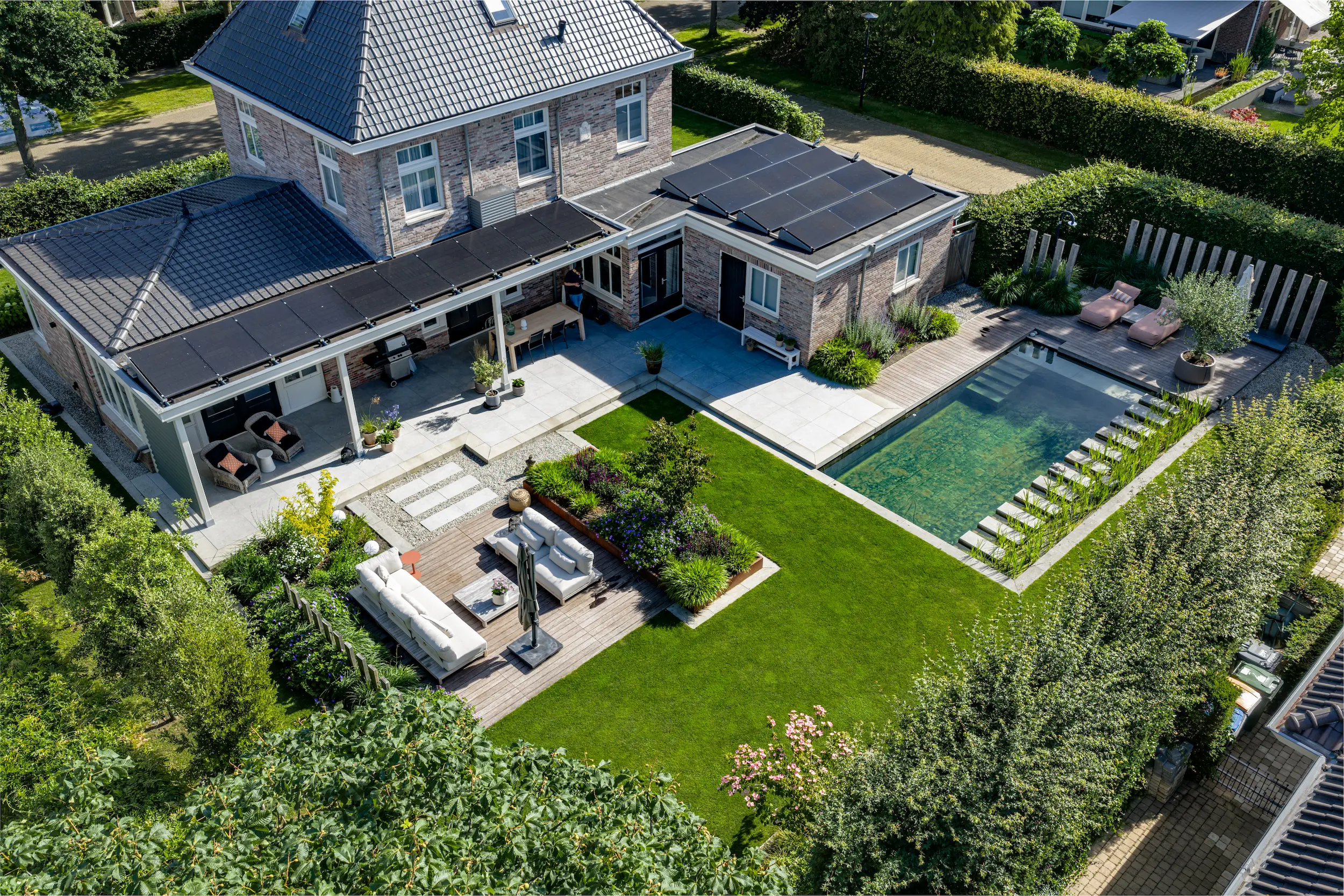 Luchtopname van villa met zonnepanelen, overkapping, loungeset op vlonder, strak gazon en natuurlijke zwemvijver met houten terras.