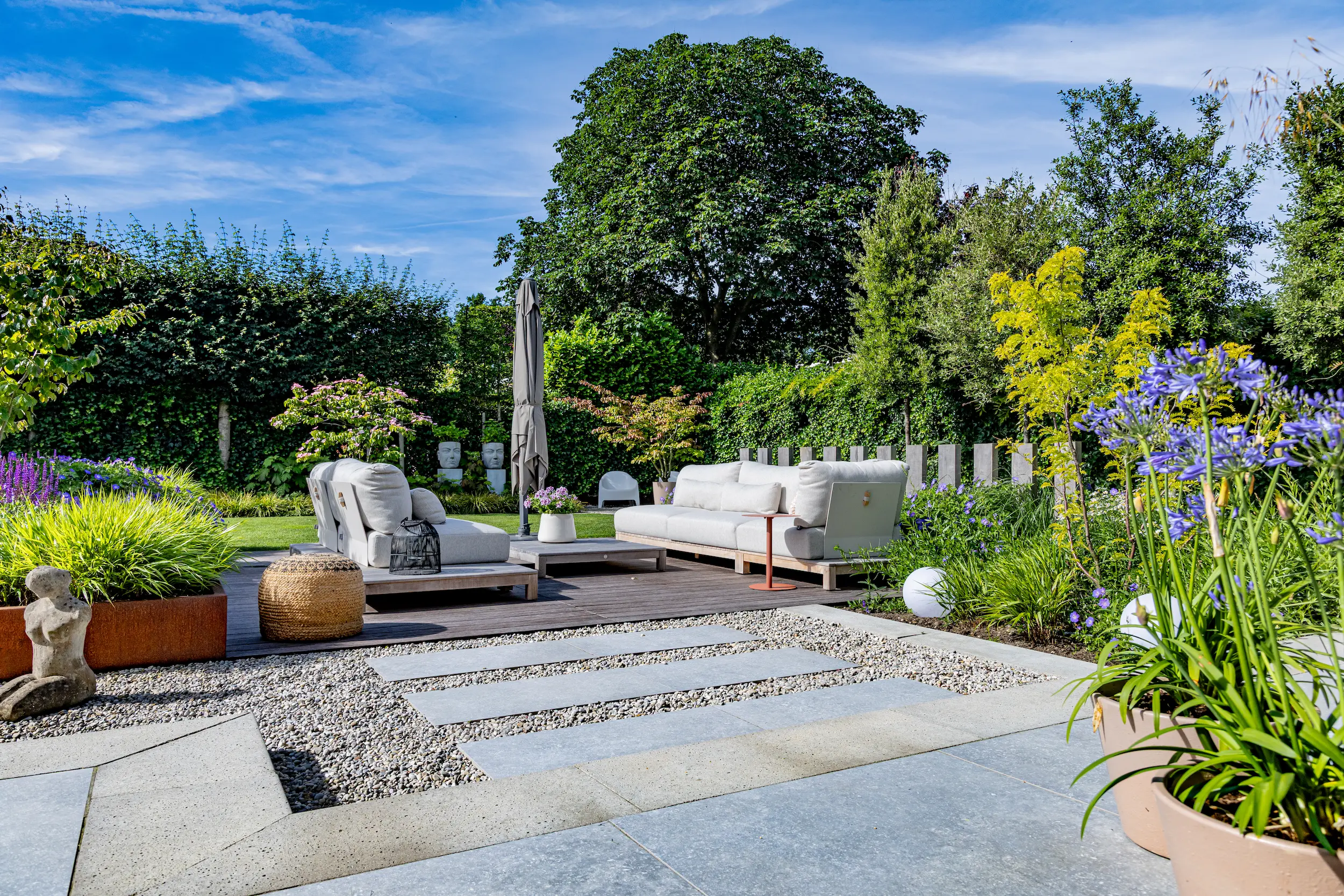Tuin met houten vlonder, witte loungebanken, parasol, grindpad met tegels, blauwe agapanthus in pot en hoge hagen.