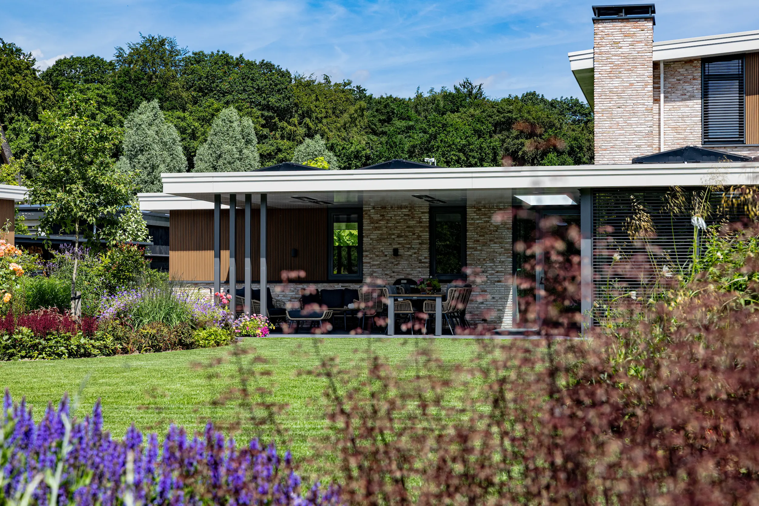Moderne villa met plat dak, overdekt terras met loungeset en eettafel, gazon en bloeiende borders op de voorgrond.