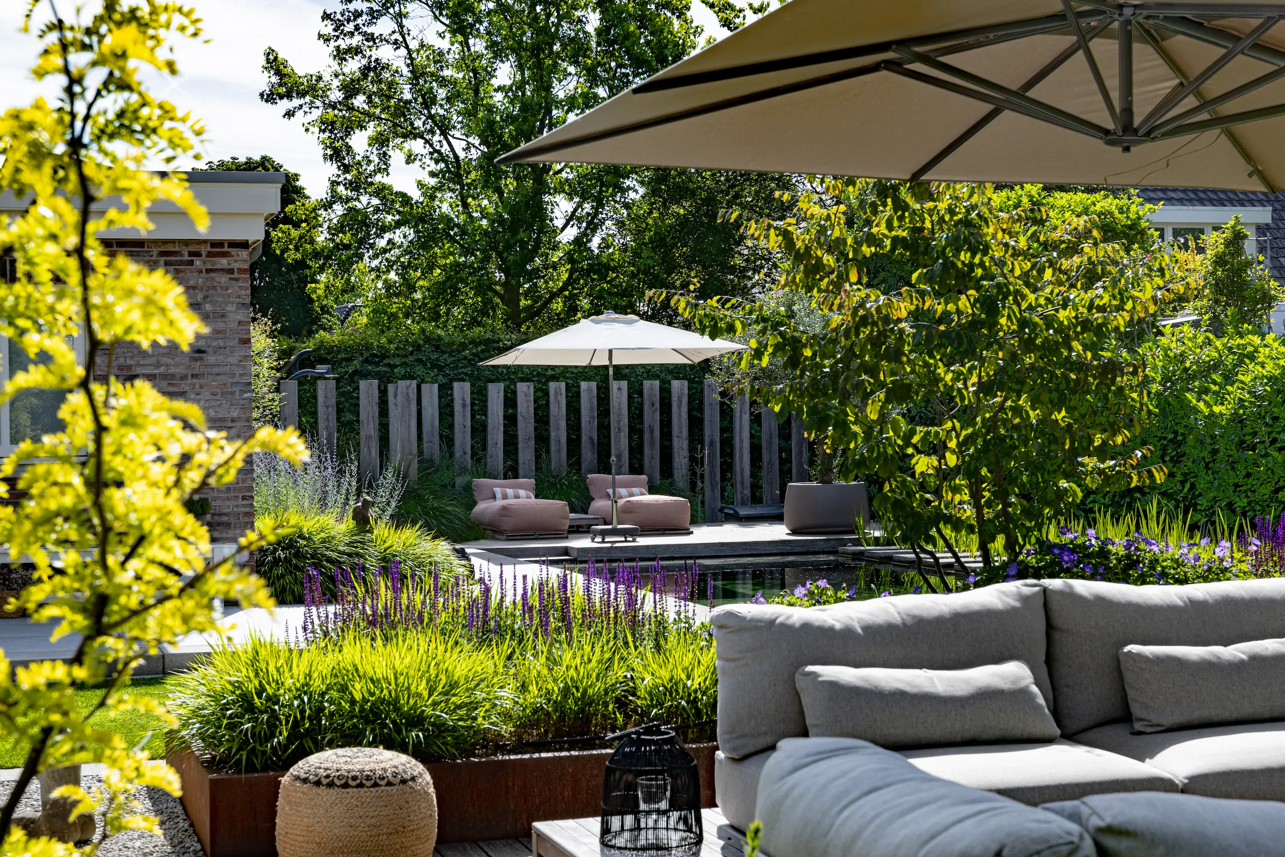 Tuin met grijze loungebank onder parasol, paarse salvia, siergrassen en loungestoelen bij een vijver voor houten schutting.